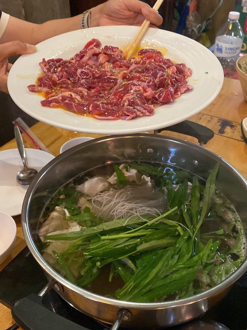 หม้อแจ่วฮ้อนกำลังเดือด มีผักบุ้งและวุ้นเส้น กับจานเนื้อสไลซ์ที่ยกอยู่เหนือหม้อ